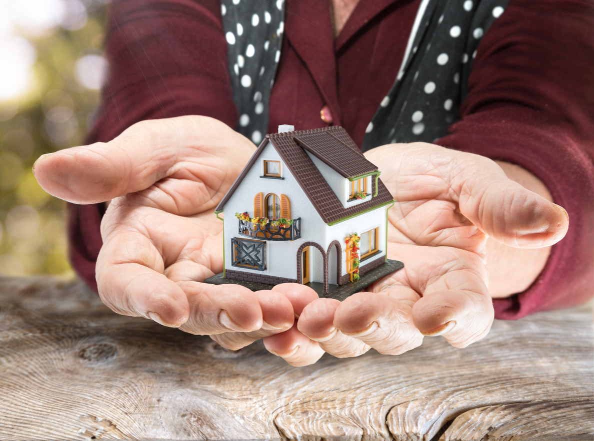 Elderly hands holding a small model house with detailed features, like windows, a door, and a balcony, against a blurred natural background. The person is wearing a polka dot top.