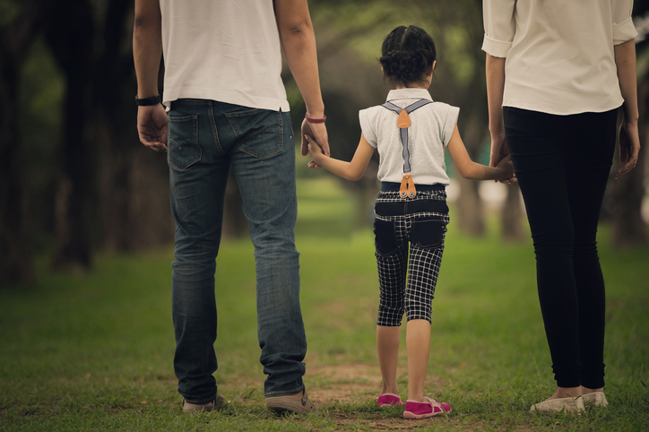 A child holding hands with two adults, one on each side. They are walking on a grassy path in a park, wearing casual clothes. The focus is on their backs, showing unity and togetherness.