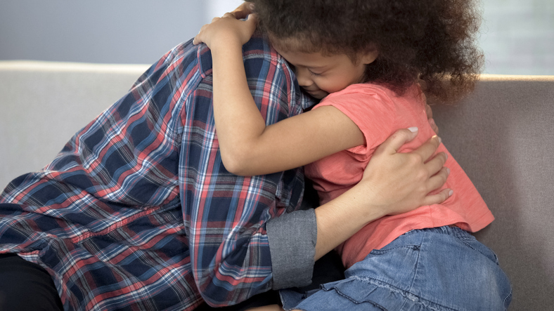 A child with curly hair hugs an adult wearing a checkered shirt on a couch. The child appears to be comforting the adult, who has their face turned away. The setting is a softly lit indoor space.