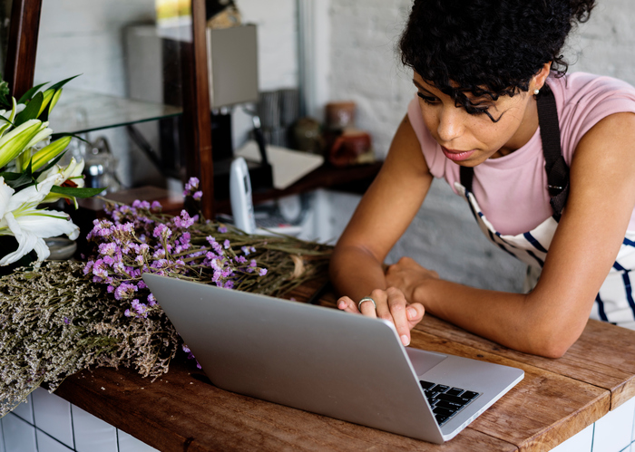 A person wearing an apron leans over a laptop on a wooden counter, surrounded by bundles of flowers, including white lilies and purple blossoms. The setting suggests a floral shop or small business environment.