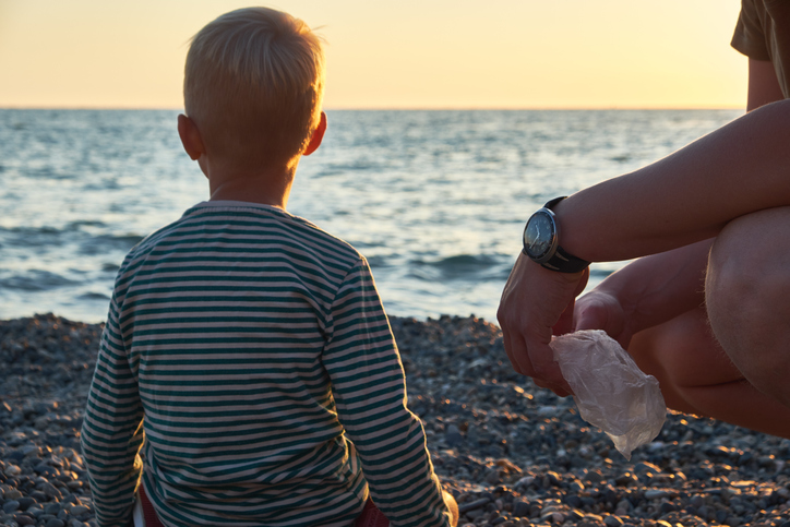 A child in a striped shirt sits on a pebble beach, facing the calm sea during sunset. An adult beside them, wearing a watch, holds a plastic bag. The sky is a warm golden hue, creating a peaceful scene.