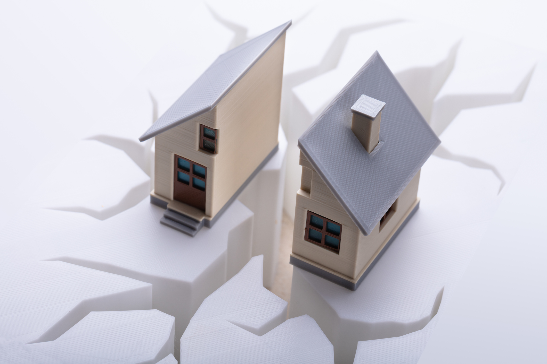Two model houses sit on opposite sides of a large, jagged crack on a white surface, symbolizing separation or disaster. The houses are detailed with grey roofs and blue windows, emphasizing the divide between them.