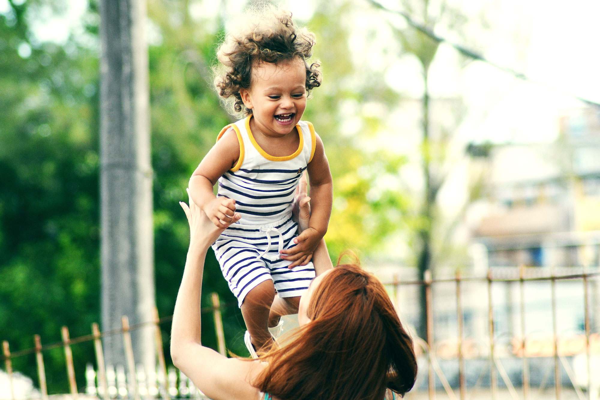 A smiling child wearing a striped dress is being lifted into the air by an adult whose face is not visible. The background features a blurred outdoor setting with trees and some buildings.