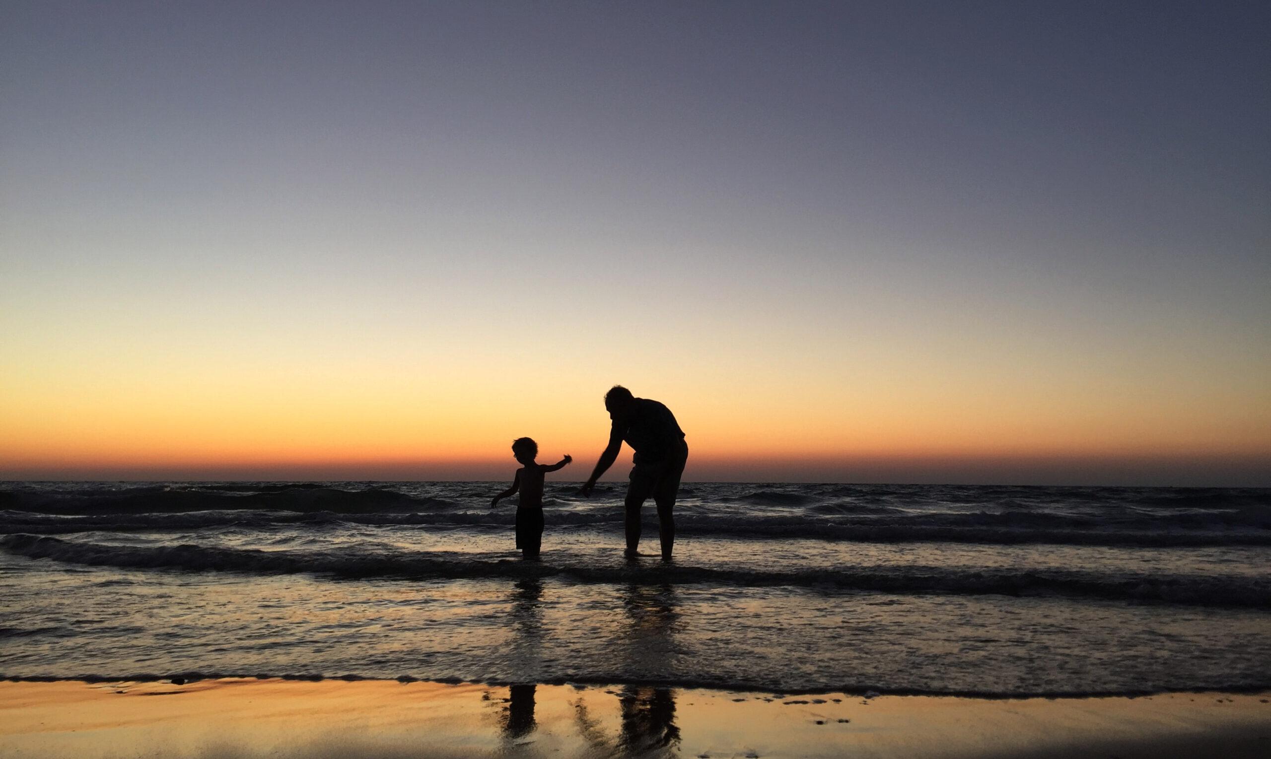 Silhouette of an adult and child holding hands at the beach during sunset, with gentle waves and a glowing sky in shades of orange and blue. Their reflection is visible on the wet sand.