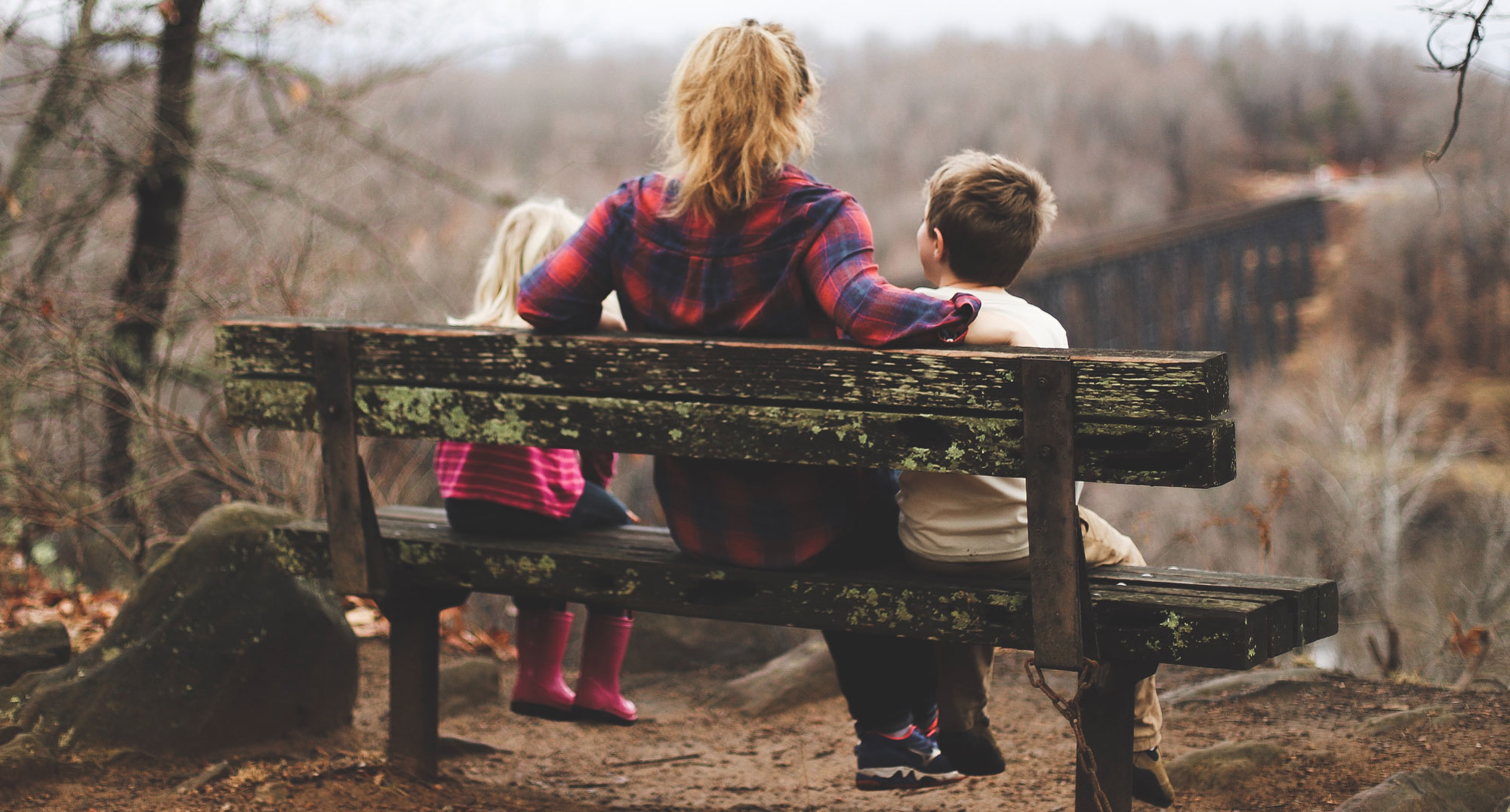 A woman and two children sit on a weathered wooden bench, overlooking a forested landscape in autumn. The woman has one arm around the boy beside her. The trees are bare, and an old railway bridge is visible in the background.