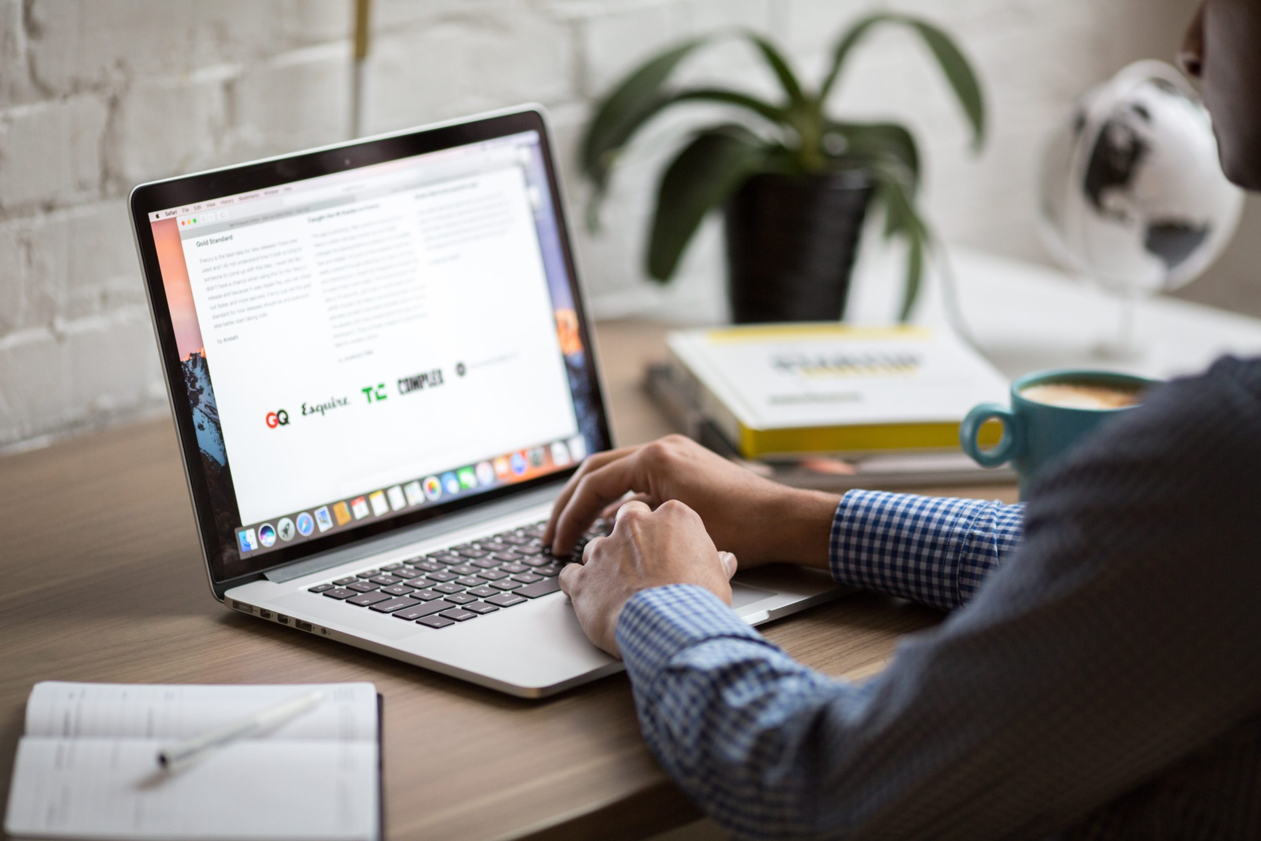 Person typing on a laptop at a wooden desk, with a potted plant, a notebook, a globe, and a cup beside them. The person wears a blue checkered shirt. The laptop screen displays a paragraph of text with some bold headings.