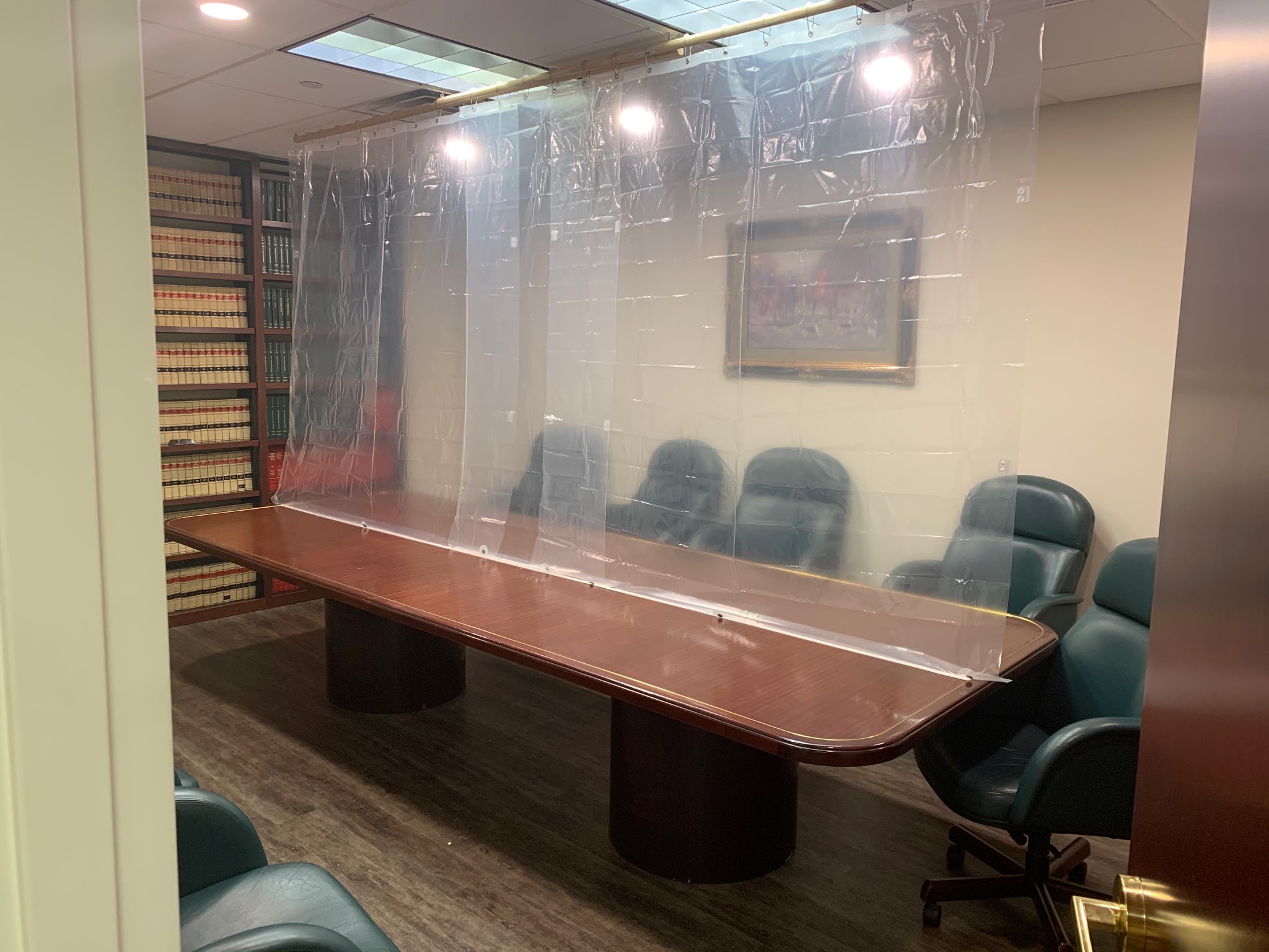 A conference room with a long wooden table surrounded by green chairs. A transparent plastic sheet divides the table. Bookshelves with many books and a framed picture are in the background.