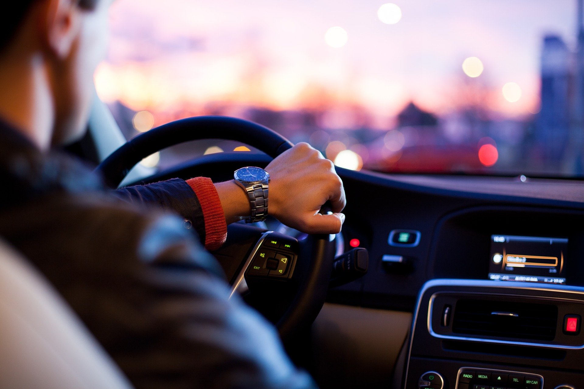 A person wearing a watch is driving a car during sunset. The interior shows a steering wheel and a dashboard with illuminated controls. The background displays a blurred view of city lights against a colorful sky.