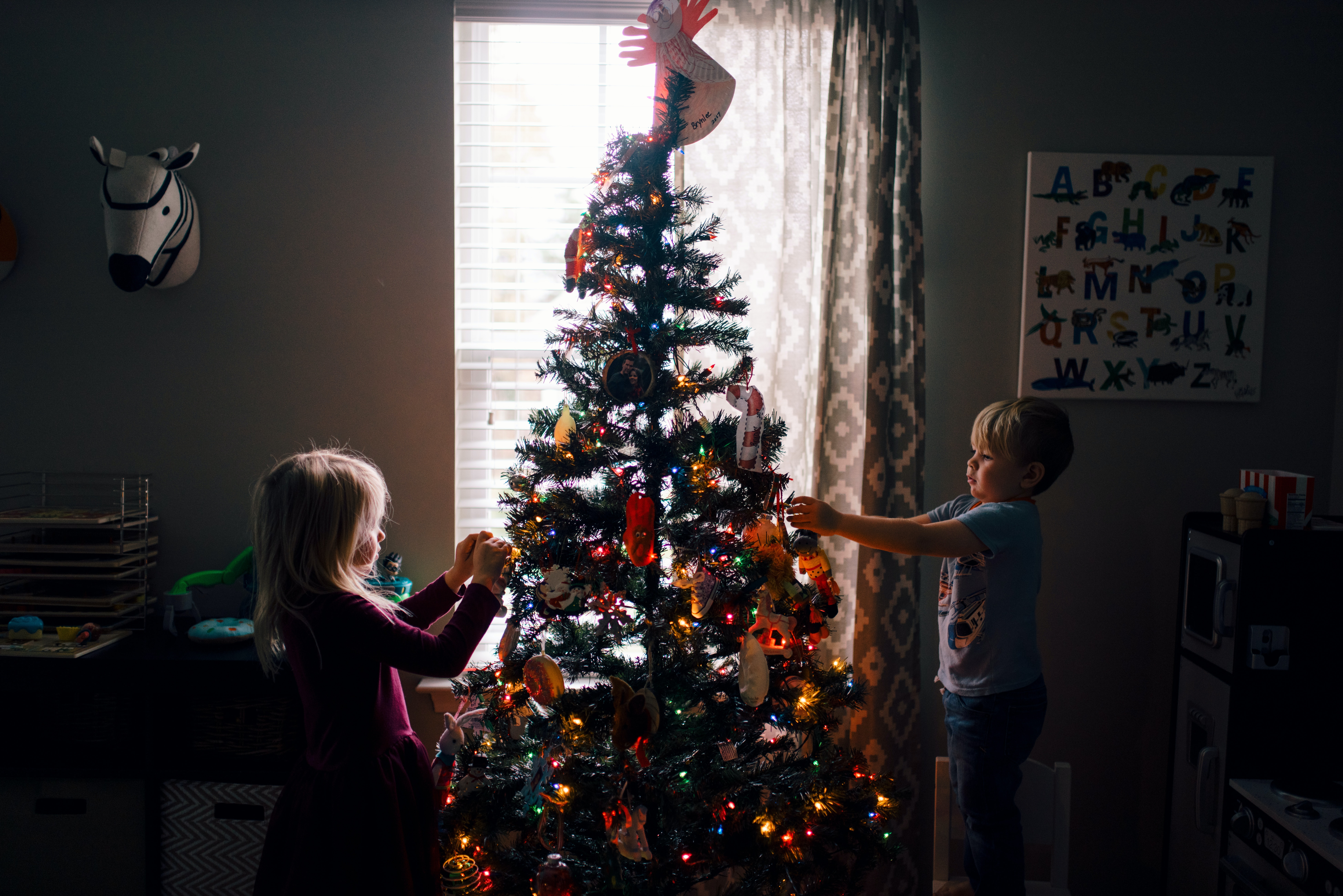 Two children are decorating a Christmas tree with colorful ornaments in a dimly lit room. Light from a window illuminates the tree, which is adorned with twinkling lights. A decorative animal head and alphabet chart are on the wall.