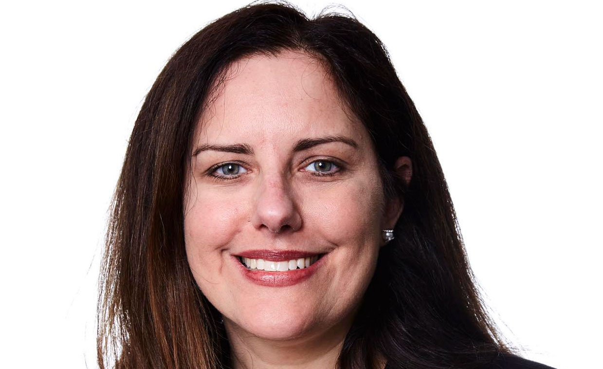 Woman with long brown hair smiling against a white background. She is wearing a black top and small earrings.