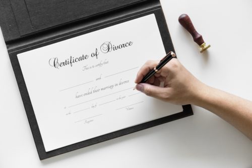 A person holding a pen is about to sign a Certificate of Divorce placed on a white surface. A seal is visible to the right of the document.