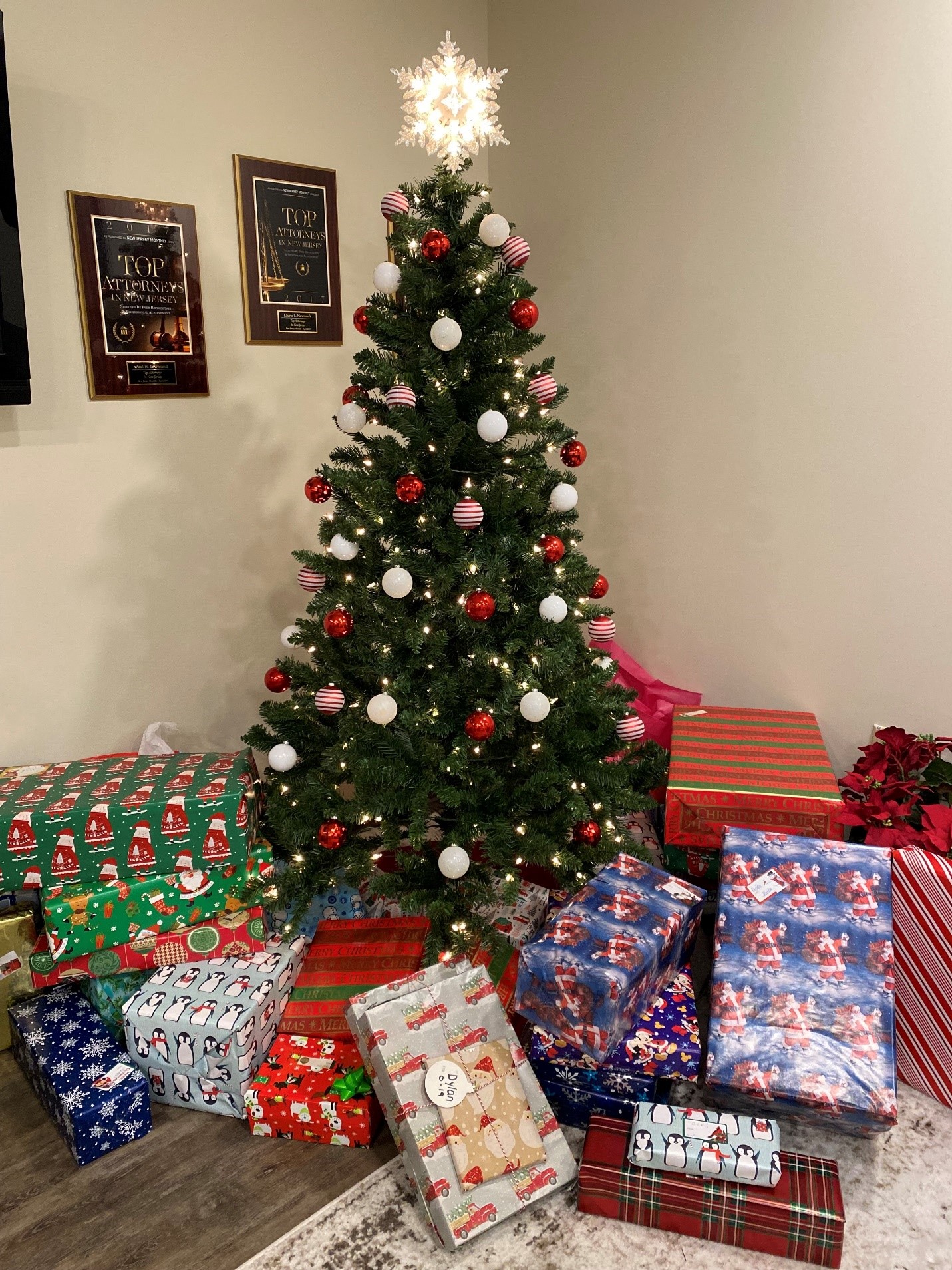 A decorated Christmas tree with white and red ornaments stands in a room. Below it are numerous wrapped gifts in colorful paper. Two award plaques are mounted on the wall. Poinsettia plants are placed on the floor to the right.