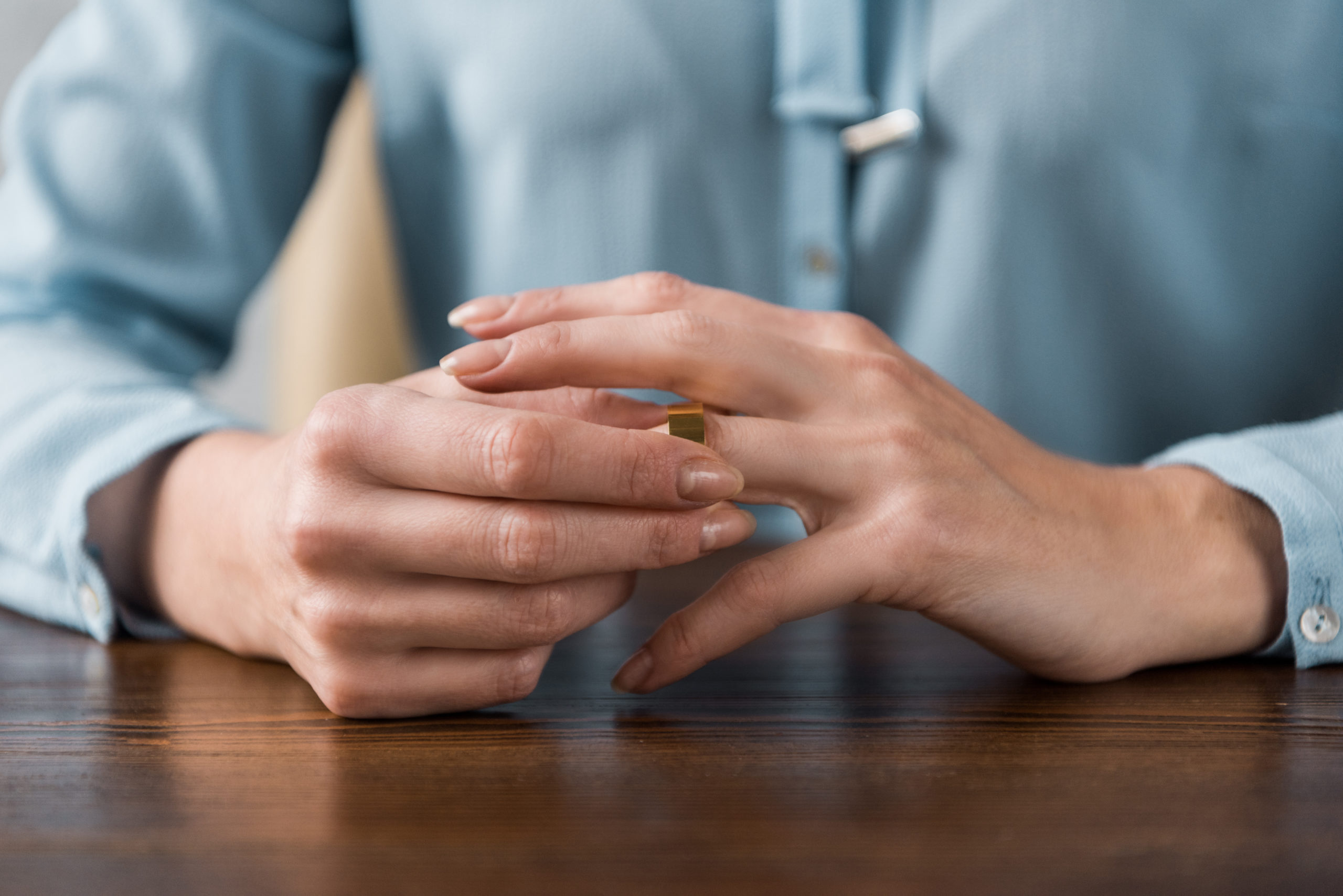 A person wearing a blue shirt is sitting at a wooden table, gently removing a gold ring from their ring finger. Their hands are centered in the image, focusing on the act of taking off the ring.