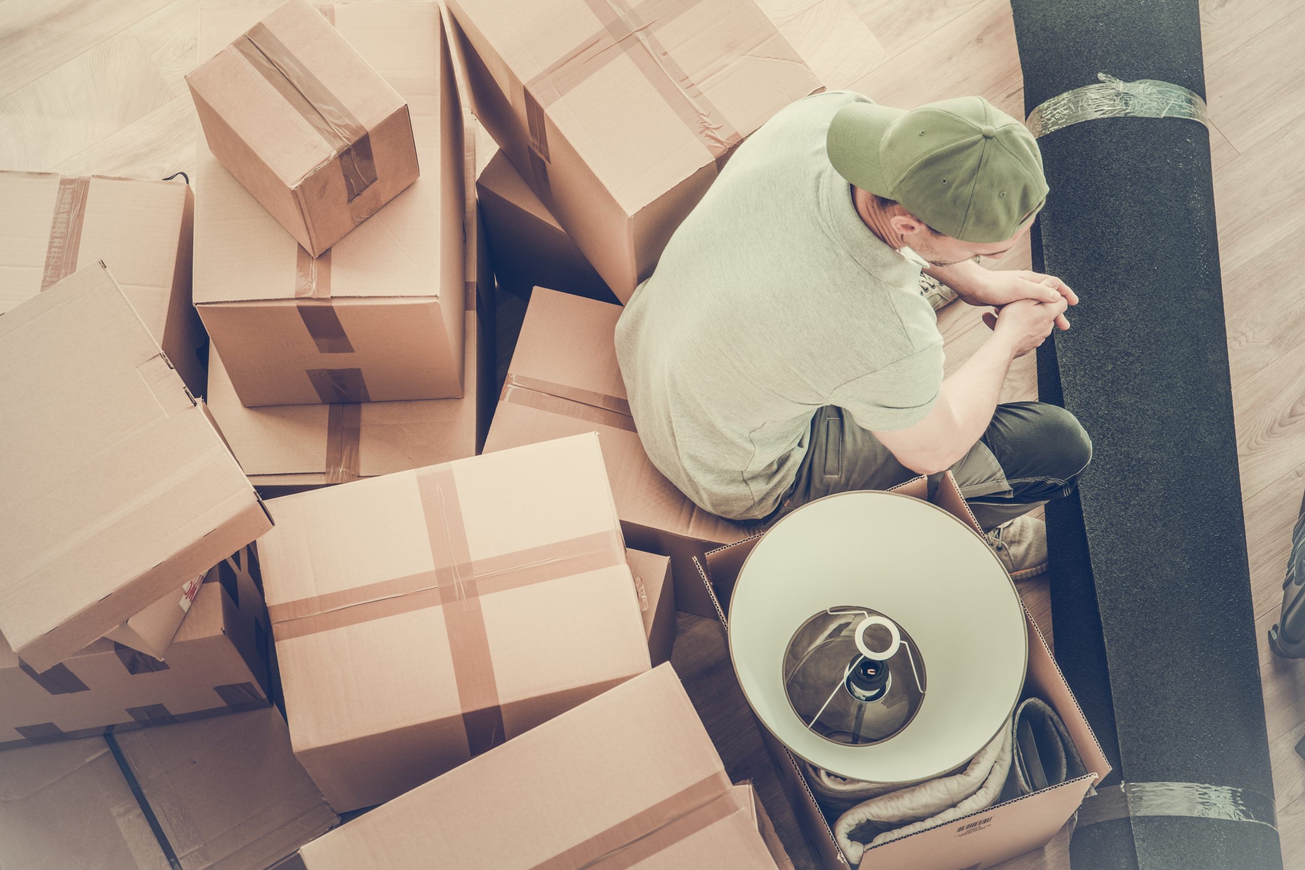 A person wearing a cap and a gray shirt sits surrounded by cardboard boxes and a carpet roll on a wooden floor. A lampshade peeks from one box, indicating a moving or packing scene.