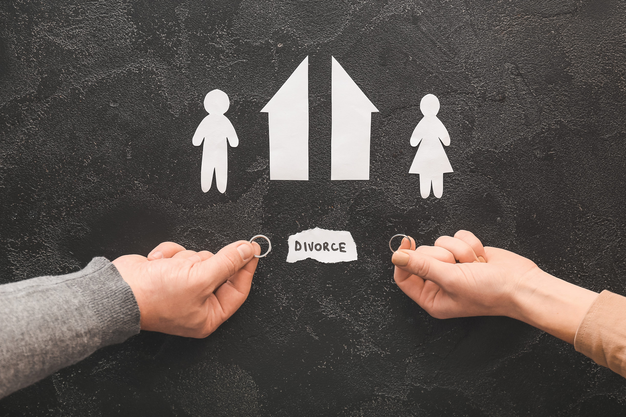 Two hands, each holding a wedding ring, reach toward cutouts of a house and two figures on a black background. The word 