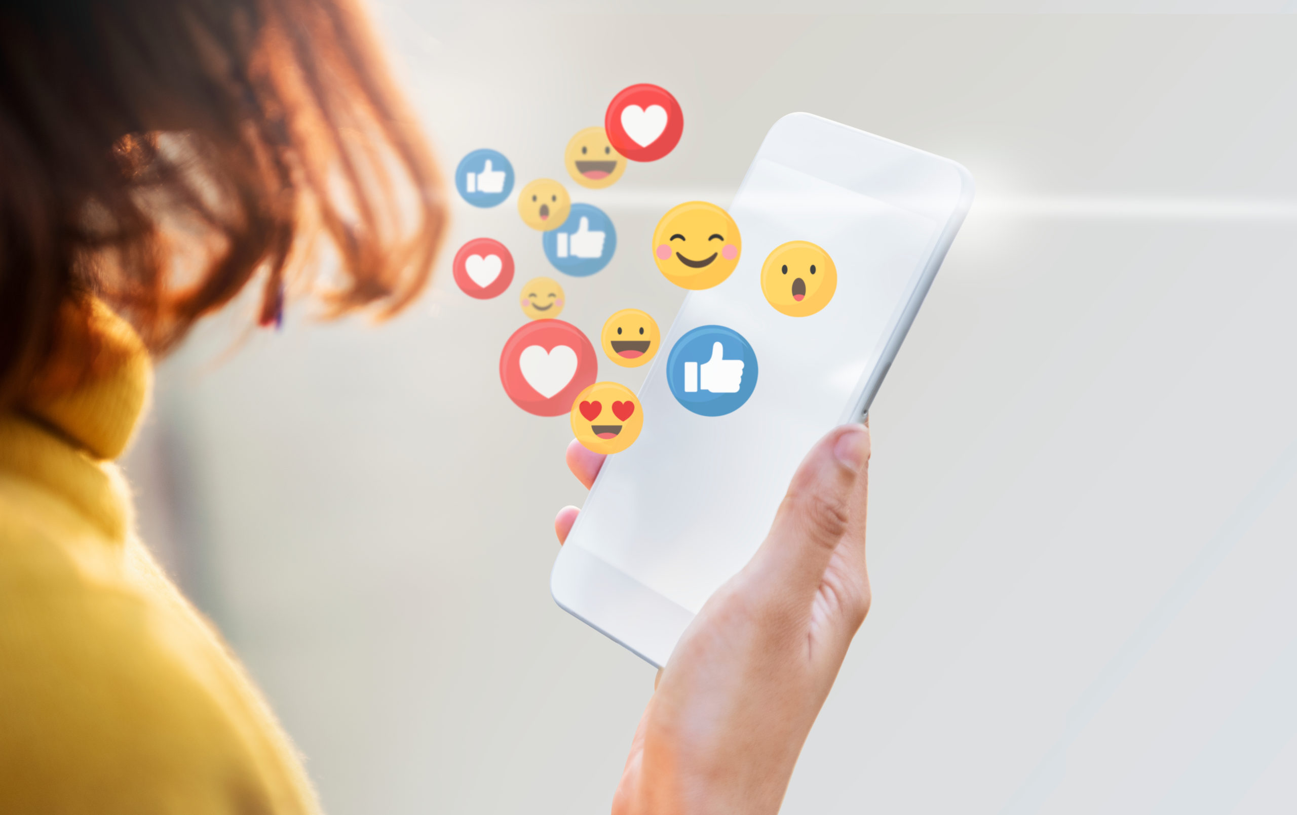 A person with shoulder-length brown hair holds a smartphone displaying various social media reaction icons, such as thumbs up, hearts, and smiley faces. The background is softly blurred, focusing attention on the phone and icons.
