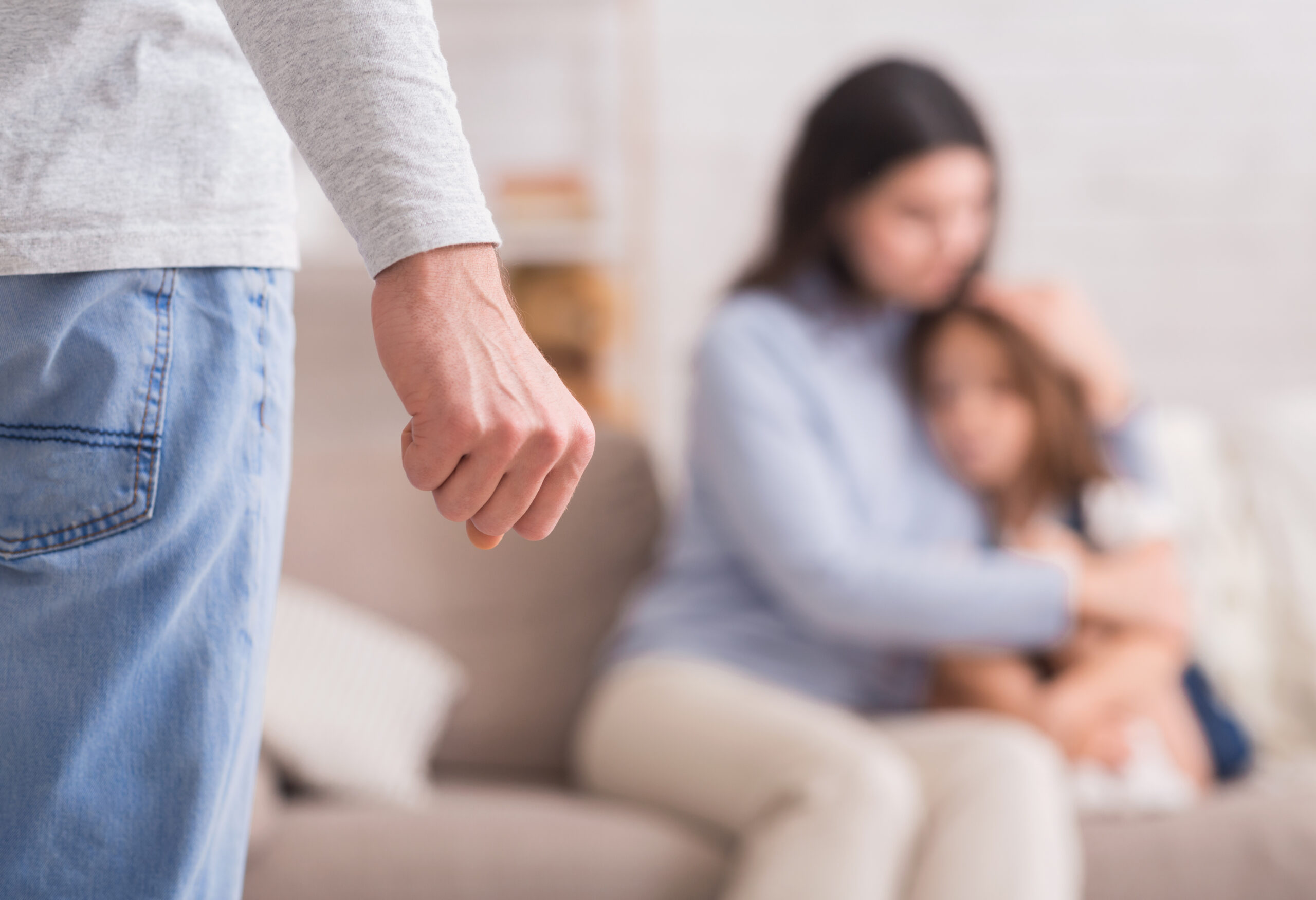 husband with clenched fist looking down at mother and child embracing in fear on a couch in family's home conveying domestic violence of wife and child abuse; Morris County Domestic Violence Lawyers concept image