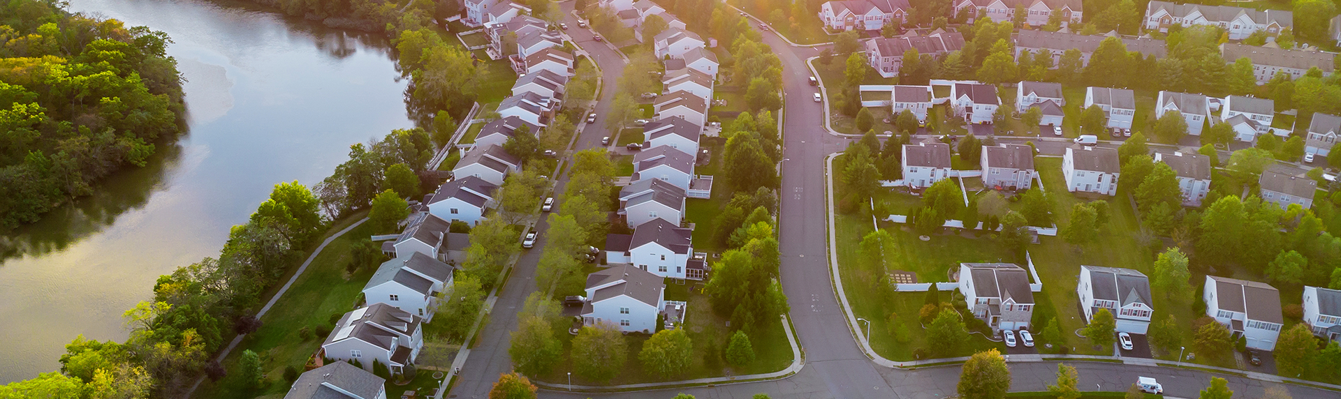 Aerial view of a suburban neighborhood with rows of houses and curved streets, reminiscent of a criminal defense lawyer's strategic path. A river runs along the top left, bordered by lush greenery. The sun casts warm light over the rooftops, creating a serene and picturesque scene.