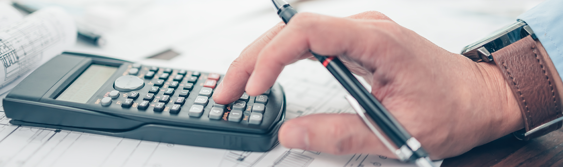 A hand wearing a watch holds a pen and presses buttons on a calculator. The calculator, surrounded by papers and architectural blueprints, suggests meticulous analysis akin to how a criminal defense lawyer meticulously reviews case details in preparation for court proceedings.