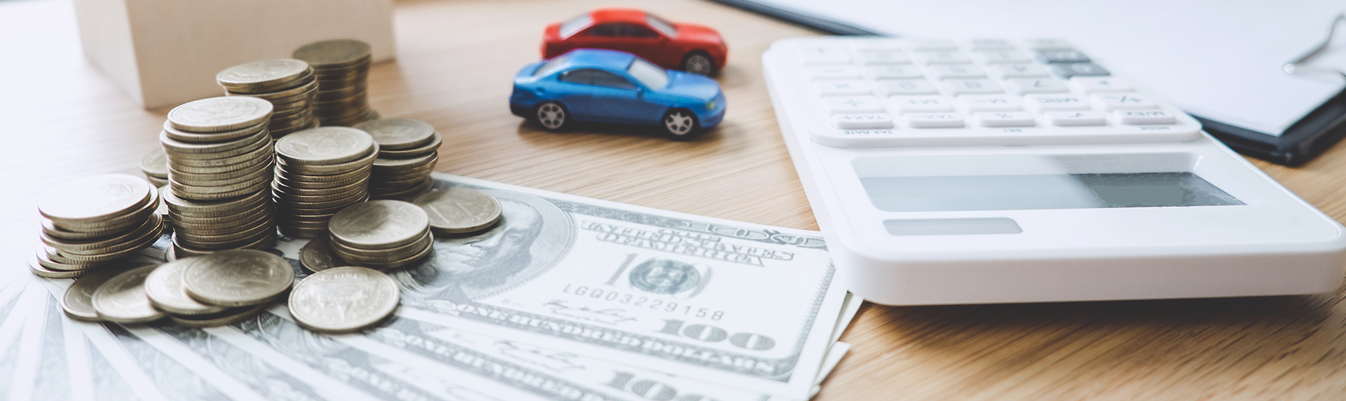 Stacks of coins and dollar bills are scattered on a wooden surface alongside a white calculator. Nearby, two small toy cars, one red and one blue, suggest financial planning related to automobiles—perhaps echoing the meticulous calculations often handled by a criminal defense lawyer.
