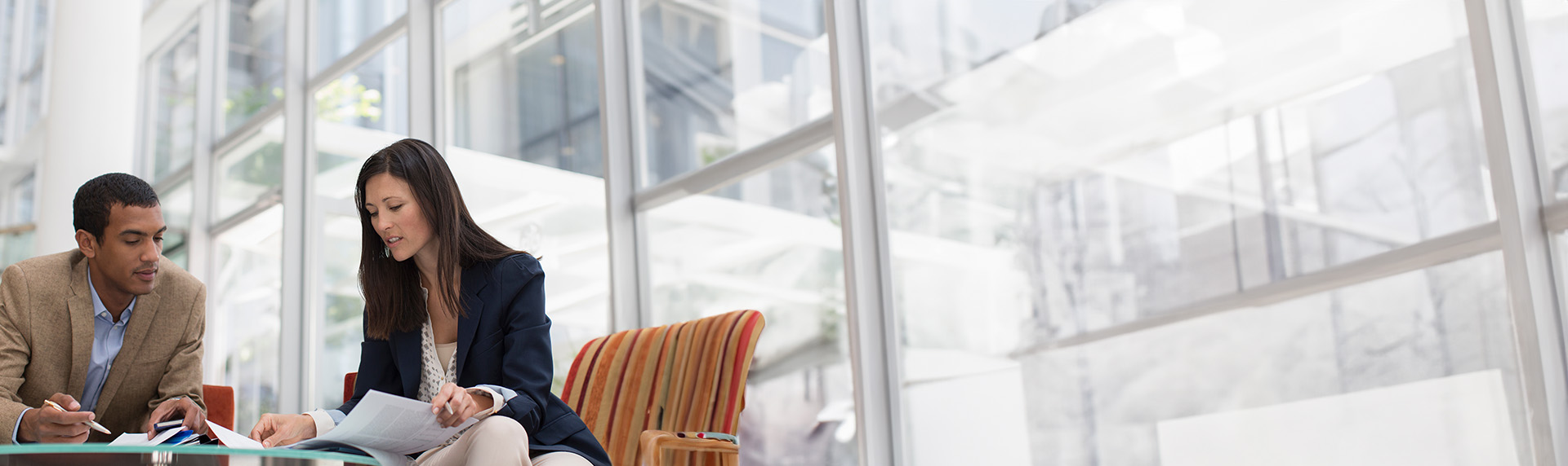 Two people sit together at a modern office table with glass walls in the background, focused on documents that lay before them. Their concentrated expressions suggest they are preparing for mediation, diving deep into details to ensure a productive discussion.