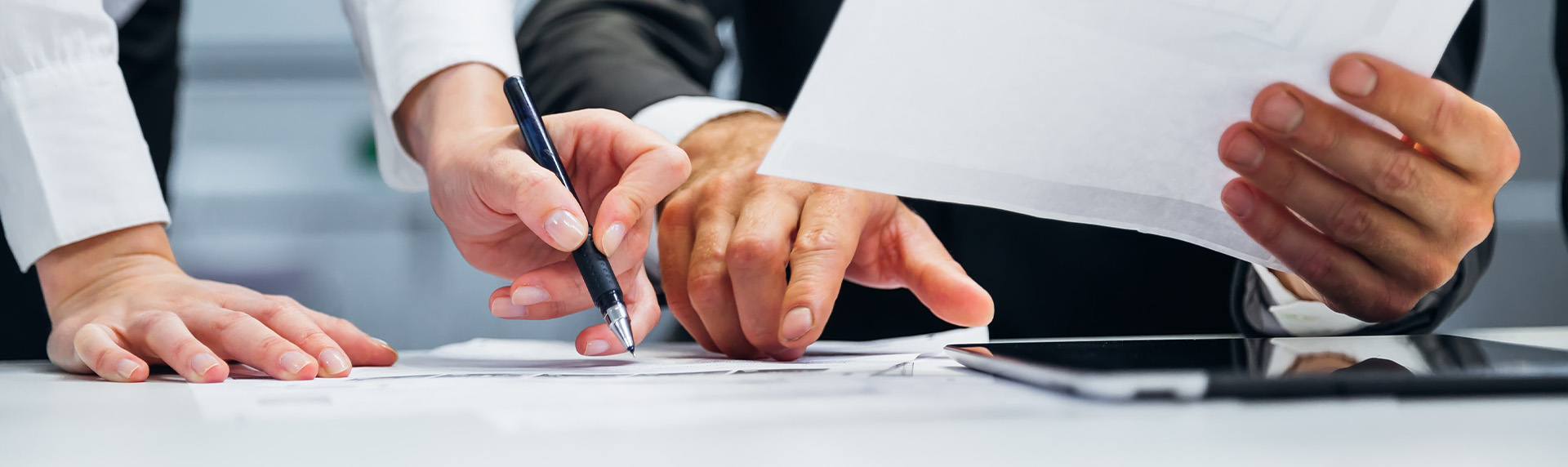 Two people reviewing documents on a table in what seems like a professional office setting. One holds a pen, pointing at the papers, while the other gestures towards a sheet. Nearby, a tablet sits ready to assist with addressing potential issues related to divorce mediation.