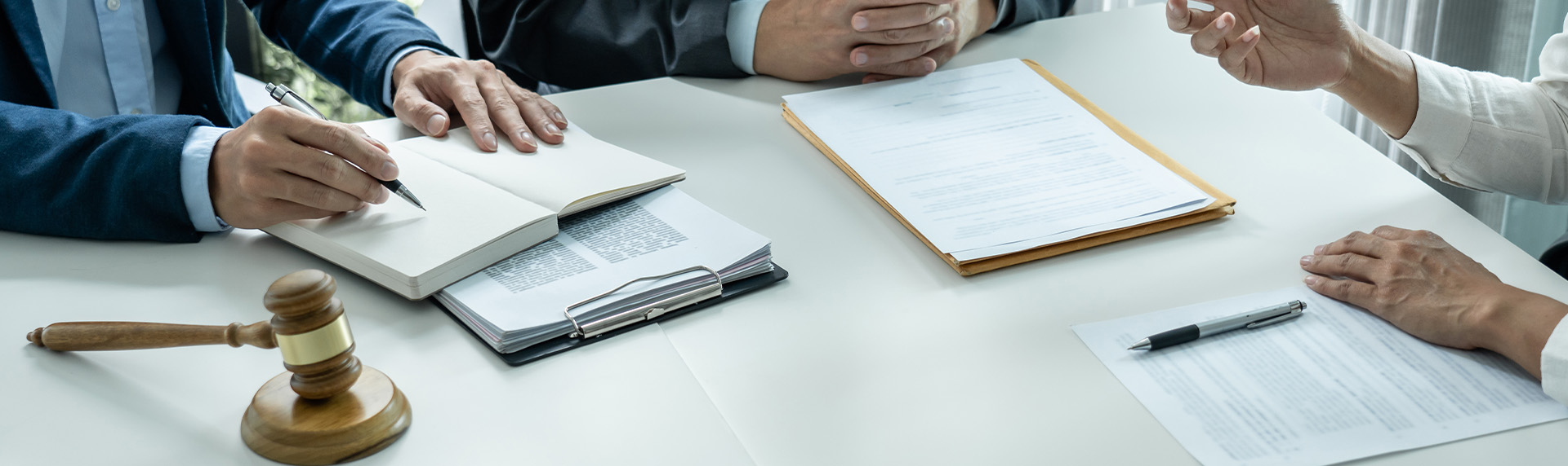 A group of people in formal attire are sitting at a table for a mediation session. Among the papers, clipboard, pen, and wooden gavel on the table, one person writes in an open notebook while another gestures, perhaps addressing a frequently asked question.