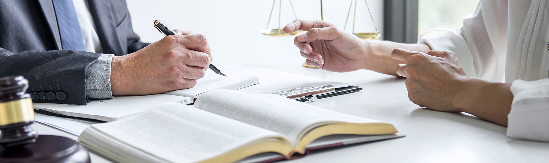 A lawyer in a suit writes intently in a notebook while consulting with a client across the desk, discussing whether mediation is right for them. An open legal book, scales of justice, and gavel adorn the table, embodying the essence of law and legal proceedings.
