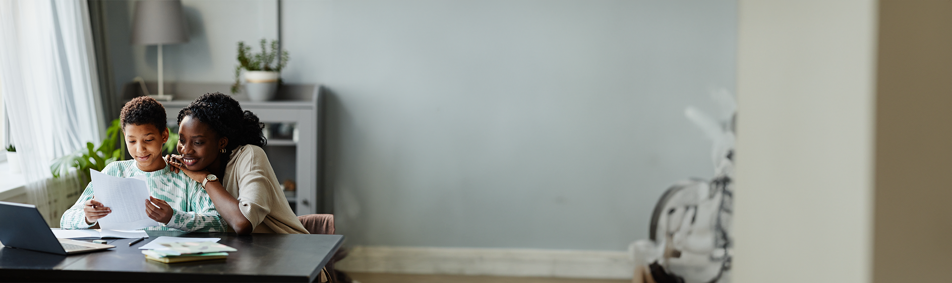 A mother and son sit at a table, smiling as they read a paper about a Criminal Defense Lawyer together. A laptop and books are on the table. The room is softly lit with a gray wall and a plant in the background, creating a cozy atmosphere.