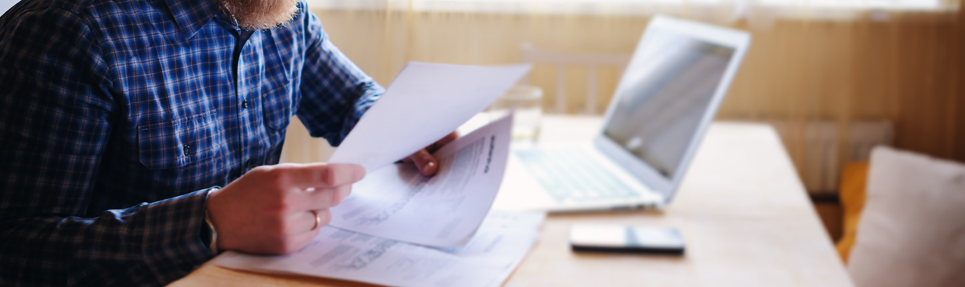 A person in a plaid shirt sits at a wooden table, meticulously reviewing papers with both hands. An open laptop and smartphone are nearby, highlighting the tools of a dedicated criminal defense lawyer. The background reveals a well-lit room with an inviting window view.