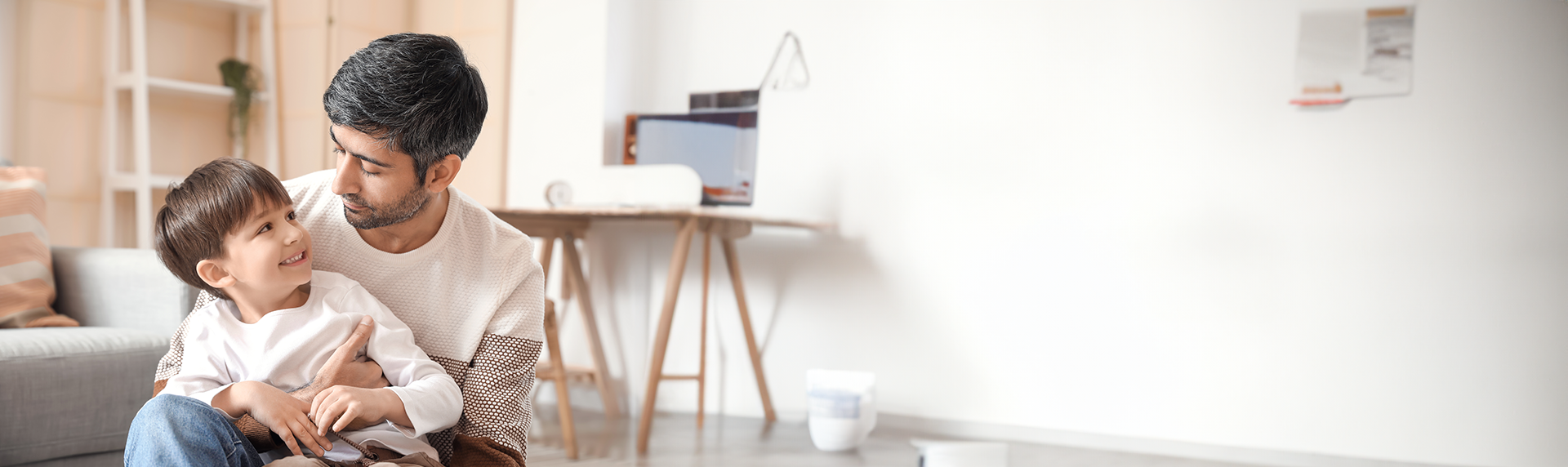 A father, a dedicated criminal defense lawyer, sits on a couch in a modern, bright living room, lovingly embracing his young son who is smiling up at him. The background features a minimalist desk and decor, creating a cozy, warm atmosphere.