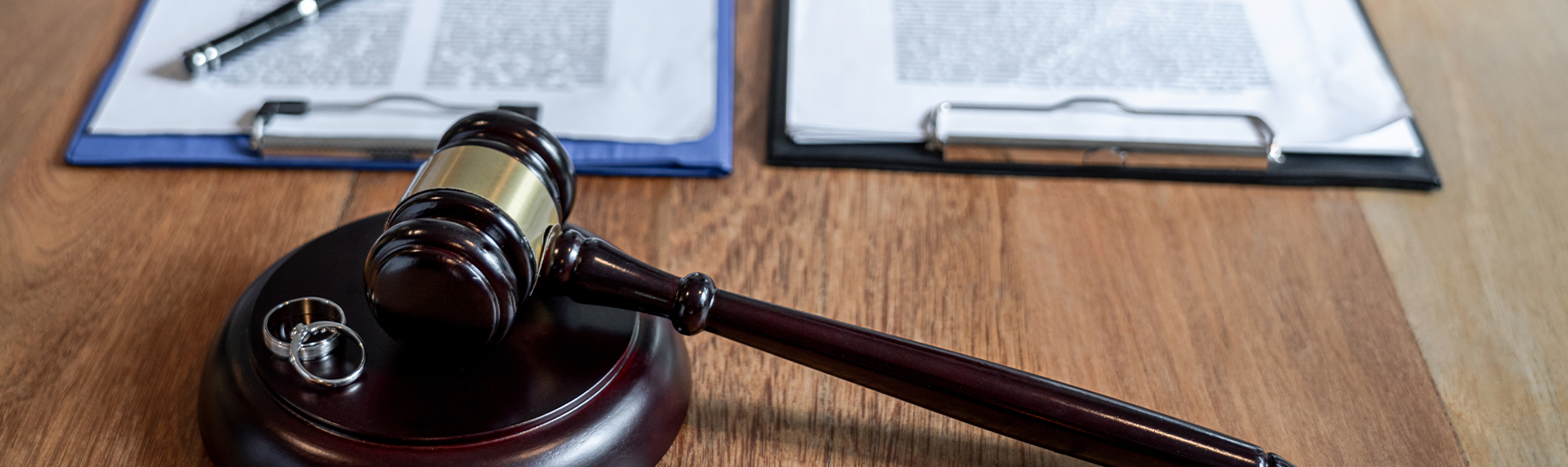 A judge's gavel and two rings rest on a wooden table, symbolizing divorce proceedings. In the background, two clipboards with documents are visible, one with a pen poised on it—echoing the meticulous preparation akin to that of a criminal defense lawyer.