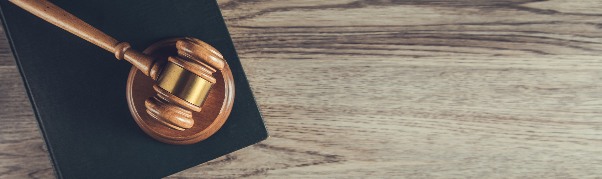 A wooden gavel rests on a sound block atop a dark book, often seen in a criminal defense lawyer's office, placed on a light wooden surface. The image is taken from an overhead perspective, with the textured wood background taking up the right side.