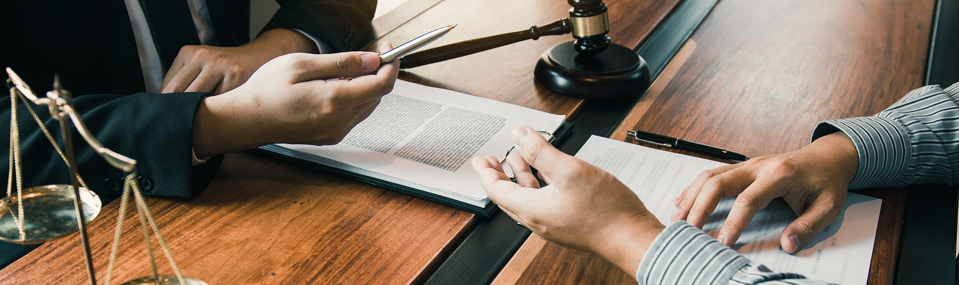 Two individuals sit at a wooden table, engaged in a discussion. One holds a pen over a document as the other listens intently. A gavel rests nearby, underscoring the legal context. Nearby, the scales of justice and elements of a mediation team enhance the scene's authoritative ambiance.