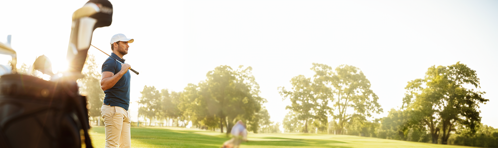 A golfer wearing a white cap and blue shirt stands on a sunny golf course, his stance as focused as a criminal defense lawyer preparing for trial. The foreground features a blurred golf bag, while the background showcases lush green grass and trees under a clear sky.