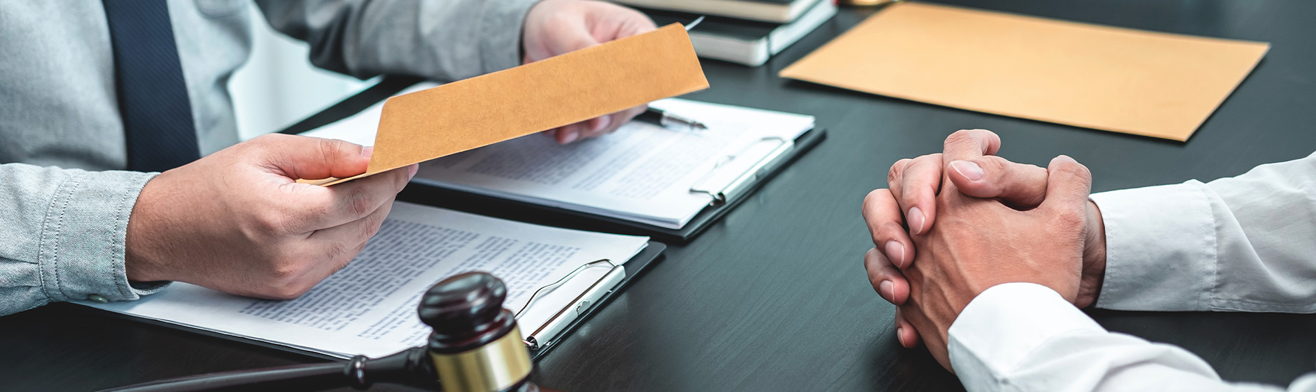 Two people are seated at a table with documents on clipboards. One, possibly a prenuptial agreement attorney, holds a brown envelope, while the other has their hands clasped. A gavel is visible on the tabletop, suggesting a legal or formal setting.
