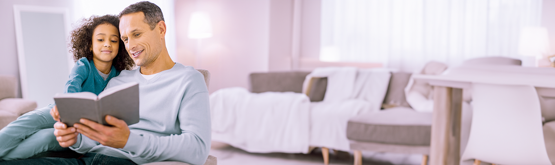 A man, a criminal defense lawyer by day, and a child sit together in a cozy, softly lit living room. The man holds a book and reads aloud, while the child leans in close, smiling. The background features a couch, pillows, and warm lighting.
