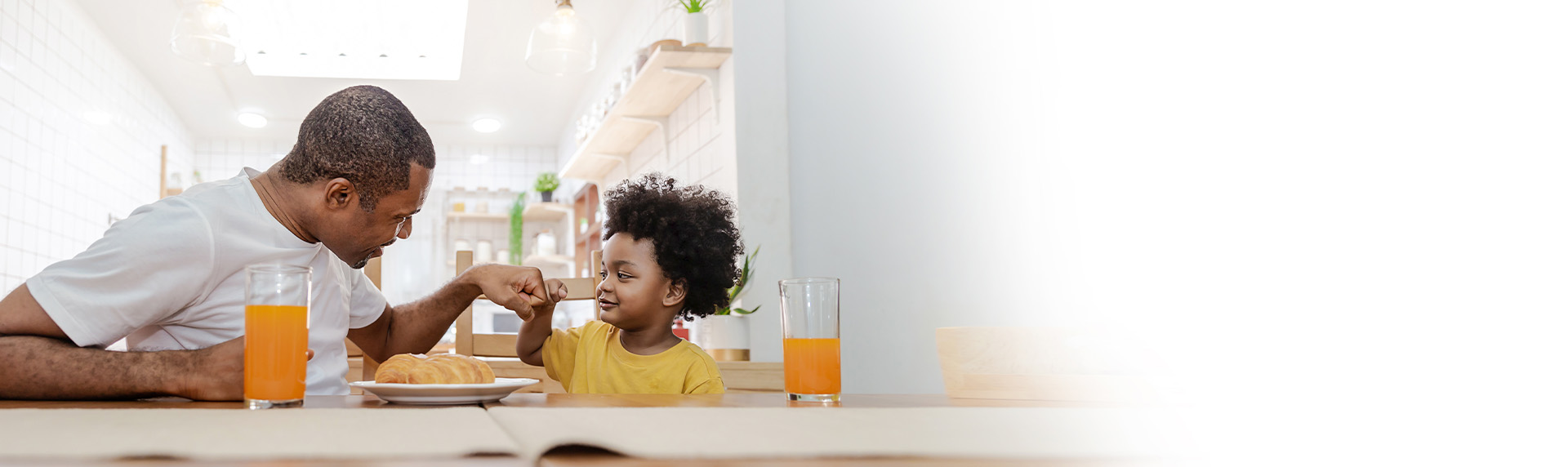 A man and a child, joyfully oblivious to the complexities of unmarried child custody, are seated at a dining table with glasses of orange juice and a plate of croissants. The man playfully interacts with the smiling child in the bright kitchen setting.