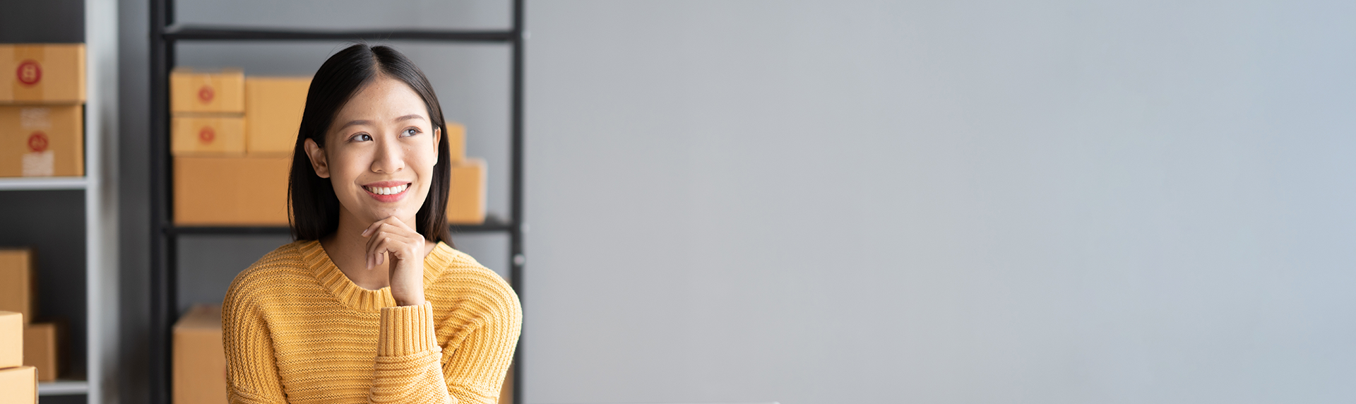 A woman in a yellow sweater sits with her hand under her chin, smiling thoughtfully like a skilled criminal defense lawyer. Behind her are shelves with cardboard boxes against a plain gray wall, suggesting a workspace or storage area.