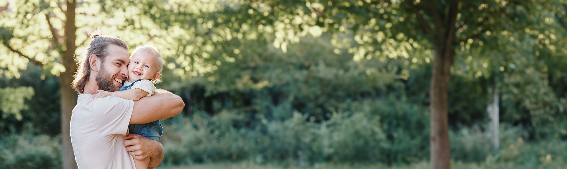 A man with a beard and pulled-back hair, who happens to be a criminal defense lawyer, joyfully holds a smiling baby in his arms. They are outdoors in a sunlit park, surrounded by lush greenery and towering trees.