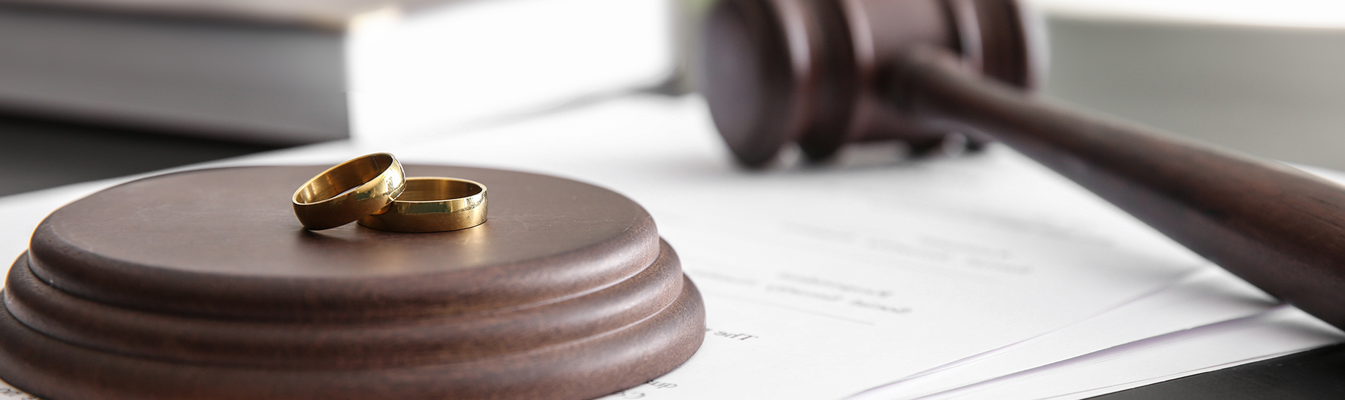 Two gold wedding rings rest on a wooden gavel stand, with a gavel nearby atop stacked documents. The scene, evoking a criminal defense lawyer's office, suggests legal proceedings possibly tied to marriage or divorce.