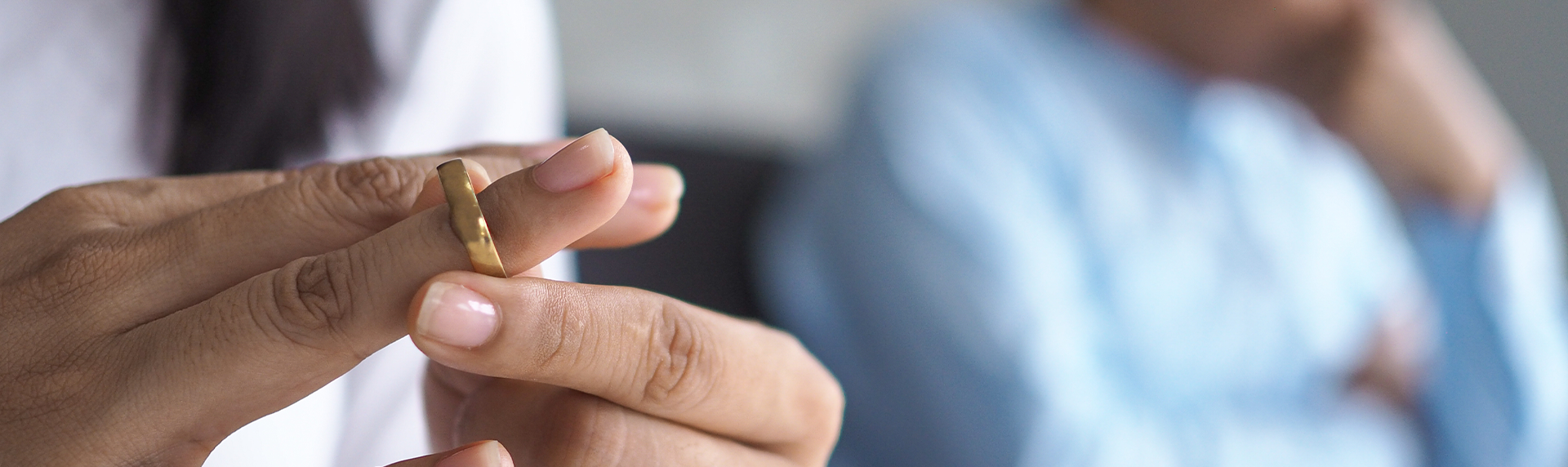 A close-up of a person sliding a gold ring off their finger, signifying tension or relationship issues. In the blurred background, another person sits wearing a blue shirt, resting their head on one hand, facing away—like conferring with a criminal defense lawyer in moments of uncertainty.