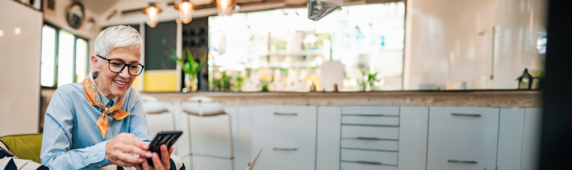 A smiling older adult with short gray hair and glasses sits in a modern kitchen, reflecting the sharp confidence of a seasoned criminal defense lawyer. They wear a blue shirt and colorful scarf as they glance at their smartphone, bright lighting casting a professional sheen over the kitchen island.