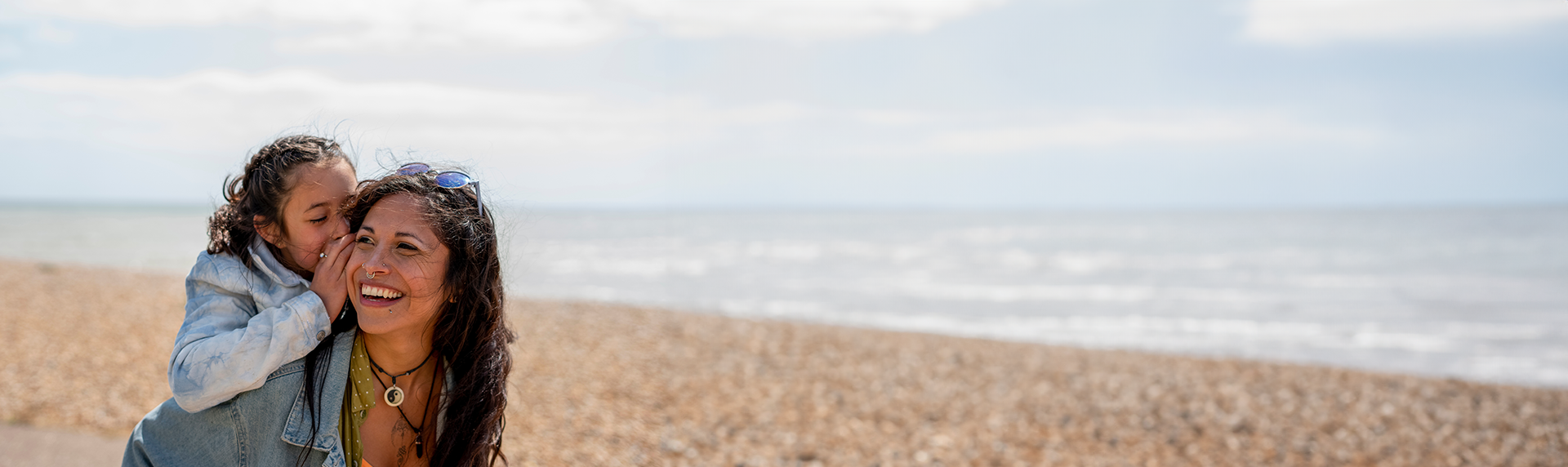 A child playfully kisses a smiling woman on the cheek by a pebbly beach, where a criminal defense lawyer might find solace after a busy day in court. The sky is partly cloudy, and the sea is visible in the background. Both appear happy and are dressed in casual clothing.
