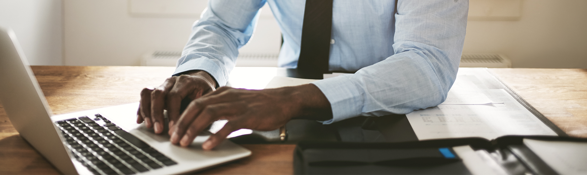 A criminal defense lawyer in a light blue shirt and tie works at a desk, diligently typing on a laptop. Various legal documents and a pen are spread out on the wooden surface as they remain focused on their case. DCPP Child Neglect Attorneys concept image