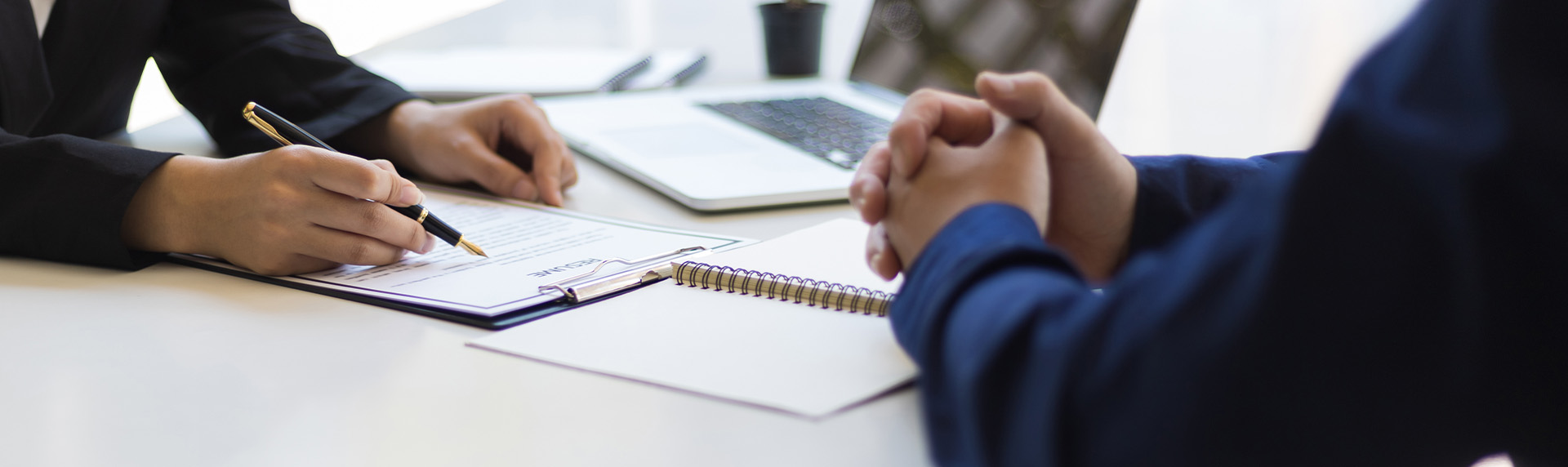 Two people are sitting at a desk, immersed in the divorce mediation process. One person is holding a pen, pointing at a document on a clipboard, while the other has their hands clasped. A laptop and notebook are also on the desk, facilitating this crucial discussion.