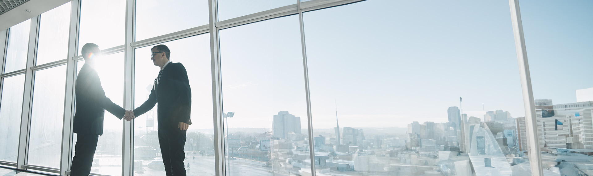 Two individuals in suits shaking hands in a modern office with large windows, the cityscape visible in the background. The scene suggests a business agreement or mediation approach, framed by skyscrapers under a clear blue sky.