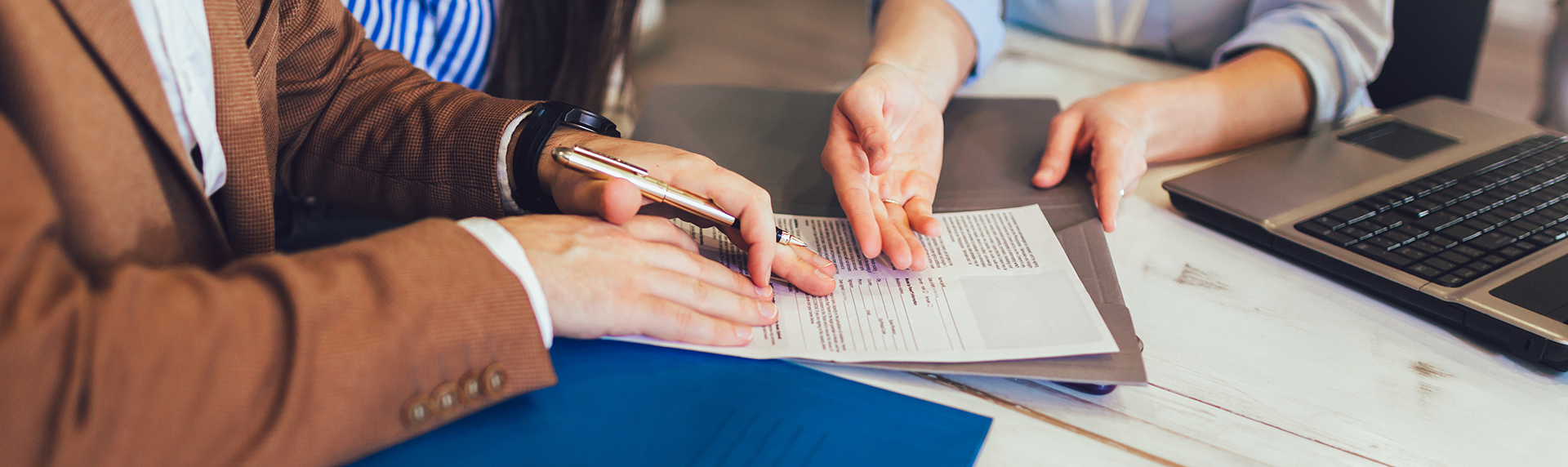 Two people are sitting at a table discussing documents. One, possibly a criminal defense lawyer, holds a pen over a document while the other points at it. A laptop and a blue folder are on the table. Both individuals' upper bodies and hands are visible.
