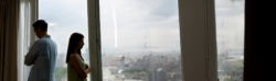 A man and woman stand back-to-back near a window, both looking upset. Overcast sky and cityscape visible outside. The woman, a seasoned criminal defense lawyer, has her arms crossed as they gaze away from each other, suggesting tension.
