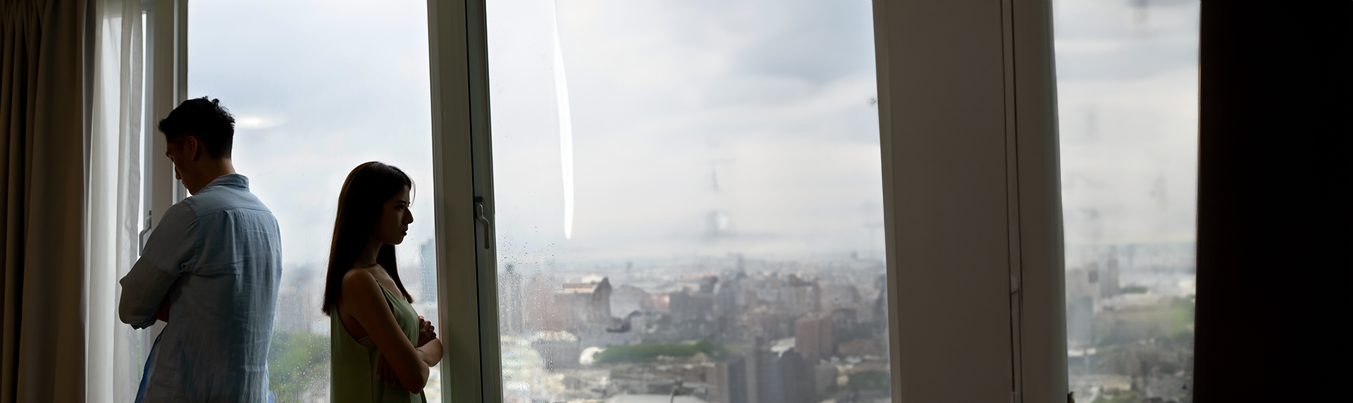 A man and woman stand back-to-back near a window, both looking upset. Overcast sky and cityscape visible outside. The woman, a seasoned criminal defense lawyer, has her arms crossed as they gaze away from each other, suggesting tension.